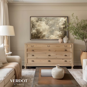 May include: A light wood dresser with five drawers and brass hardware. The dresser is in a living room with a beige wall, a painting of a cloudy sky, and a potted olive tree.