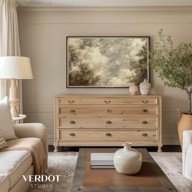 May include: A light wood dresser with five drawers and brass hardware. The dresser is in a living room with a beige wall, a painting of a cloudy sky, and a potted olive tree.