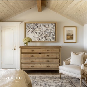 May include: A light wood dresser with four drawers and brass hardware sits in a bedroom with a white door, a beige armchair, and two framed pictures on the wall. One picture features a watercolor painting of leaves and the other picture features a landscape scene.