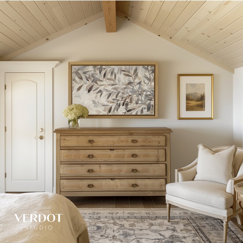 May include: A light wood dresser with four drawers and brass hardware sits in a bedroom with a white door, a beige armchair, and two framed pictures on the wall. One picture features a watercolor painting of leaves and the other picture features a landscape scene.