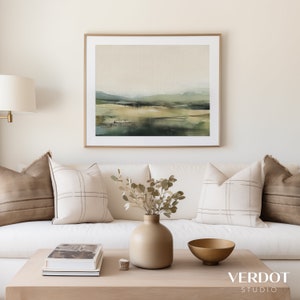 May include: A white couch with brown and white patterned pillows, a vase with dried branches, a book on a wooden coffee table, and a gold bowl. The wall behind the couch is white and features a framed landscape painting with green and brown tones.