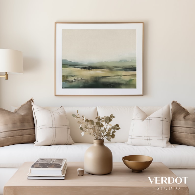 May include: A white couch with brown and white patterned pillows, a vase with dried branches, a book on a wooden coffee table, and a gold bowl. The wall behind the couch is white and features a framed landscape painting with green and brown tones.