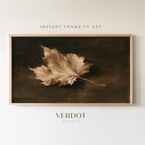 May include: A rectangular digital artwork for a television screen featuring a single dried maple leaf in shades of beige and brown against a dark, moody background. The artwork is displayed within a light-colored wooden frame. Text reads INSTANT FRAME TV ART and VERDOT STUDIO.