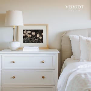 May include: White nightstand with three drawers and gold knobs. A lamp with a white shade and a white ceramic base sits on top of the nightstand. A framed print of white tulips on a dark background sits on top of the nightstand. The nightstand is next to a white bed with a beige headboard.