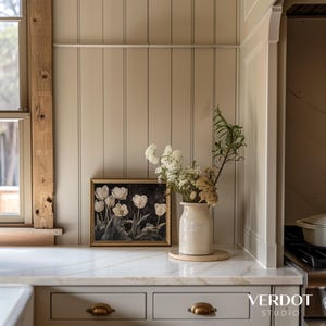 May include: A framed print of white tulips in a vase on a white countertop with a wood panel wall behind it. The vase is a cream color and has a rustic look. The countertop is made of marble.