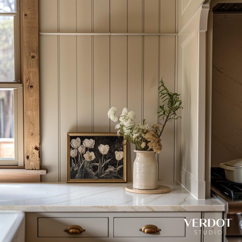 May include: A framed print of white tulips in a vase on a white countertop with a wood panel wall behind it. The vase is a cream color and has a rustic look. The countertop is made of marble.