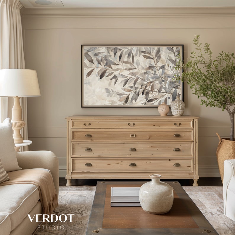 May include: A light wood dresser with five drawers and brass hardware. The dresser is against a beige wall with a framed watercolor painting of leaves above it. A small potted olive tree is on the right side of the dresser.