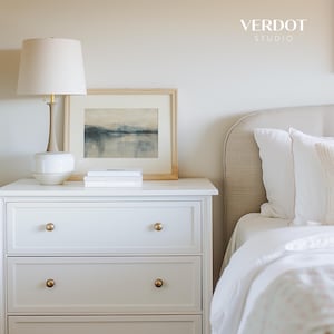 May include: White nightstand with three drawers and gold knobs. A lamp with a white shade and a white ceramic base sits on top of the nightstand. A framed watercolor painting of a landscape is on the nightstand. The nightstand is next to a white bed with a beige headboard.