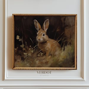 Op de afbeelding: Een ingelijst schilderij van een bruin konijn dat in hoog gras zit. Het konijn kijkt recht naar de kijker. Het schilderij is tegen een donkere achtergrond en heeft een gouden lijst.