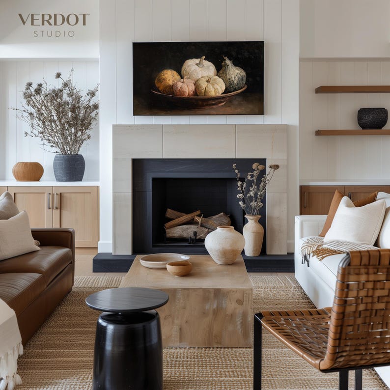 May include: A living room scene featuring a fireplace with a painting of pumpkins above it. The room has a neutral color palette with a brown leather sofa, a white sofa, and a wooden coffee table. Decorative vases and dried plants add texture.