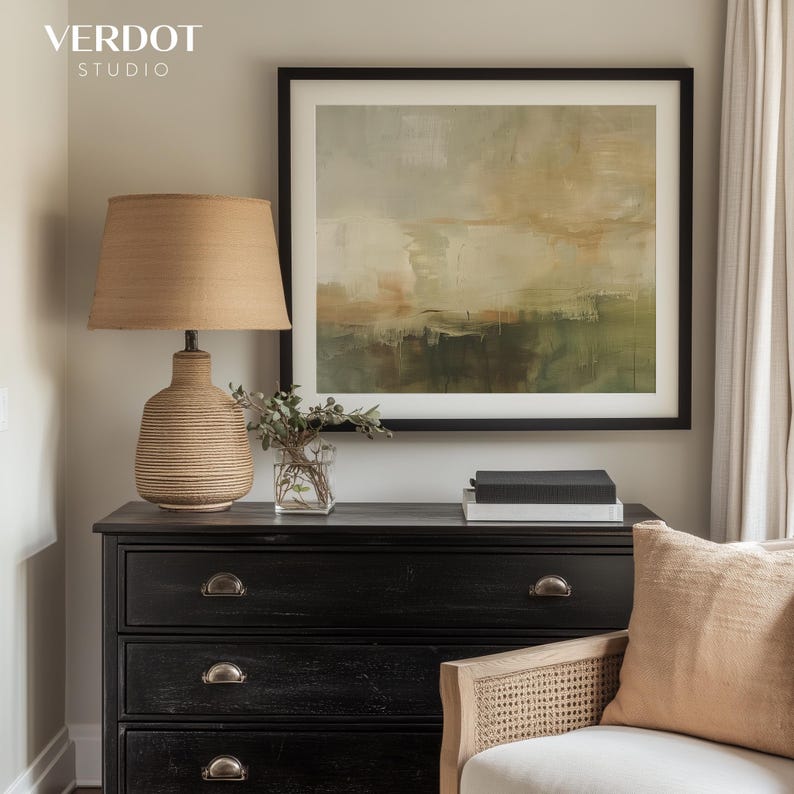 May include: A black dresser with three drawers and silver hardware. A lamp with a woven shade sits on top of the dresser. A framed abstract painting with green, brown, and white colors hangs on the wall above the dresser. A woven chair with a beige cushion sits in front of the dresser.
