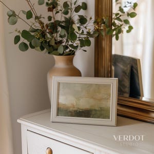 May include: A framed abstract painting with muted tones of green, brown, and yellow. The painting is in a white frame and is sitting on a white dresser with a vase of eucalyptus branches.