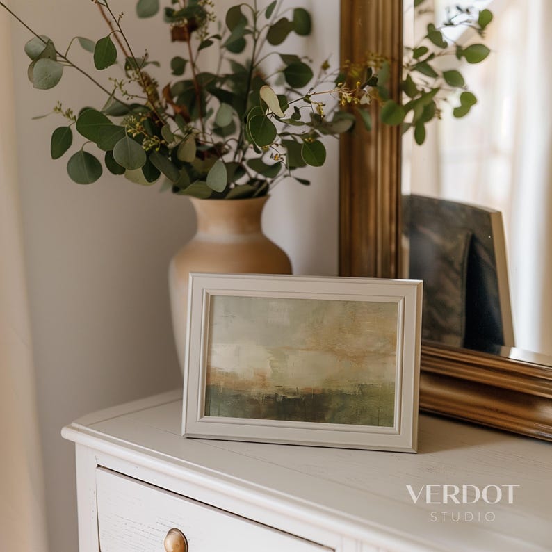 May include: A framed abstract painting with muted tones of green, brown, and yellow. The painting is in a white frame and is sitting on a white dresser with a vase of eucalyptus branches.