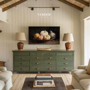 May include: A room interior featuring a green dresser with multiple drawers, two lamps with beige shades, and a painting of pumpkins. The wall behind the dresser is white, and the ceiling has exposed wooden beams. The room has a rustic, farmhouse aesthetic.