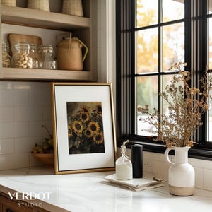 May include: A white ceramic pitcher with a brown base sits on a white countertop next to a framed print of sunflowers. A clear glass bottle with a cork stopper and two black cylindrical containers are on a white tray in front of the pitcher.