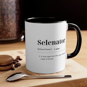 May include: White ceramic mug with a black rim and handle. The mug has the word "Selenator" printed on it, with the definition "[Selena Gomez] • noun • A loyal super fan of the cutest American singer of all time."
