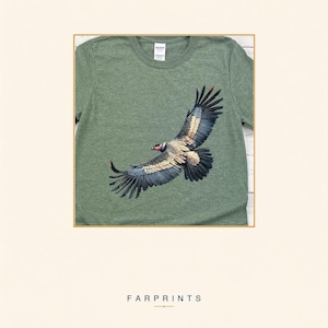 May include: A heathered olive green t-shirt featuring a detailed graphic of a condor in flight with outstretched wings. The bird displays black, white, and tan plumage with a distinct red head. The shirt has a crew neckline and a standard short-sleeve cut.