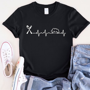 Puede incluir: Camiseta negra con un gráfico blanco de un gorro de cocinero y un tenedor y una cuchara con una línea de latido del corazón.