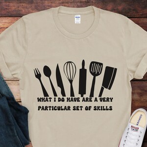 Puede incluir: Una camiseta beige con un gráfico negro de utensilios de cocina y el texto "What I do have are a very particular set of skills".