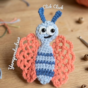 May include: A handmade crochet butterfly in light blue, with blue and white striped body, blue antennae, and coral-colored openwork wings. The butterfly has black eyes and a smiling mouth. The text "Click Clack" is above the butterfly, and "Yarnproductsua" is on the left side.