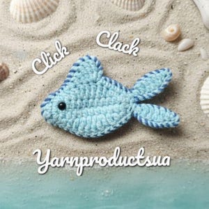 Puede incluir: Un pez de ganchillo azul claro con ribete azul oscuro, sobre arena con conchas marinas. El pez tiene un ojo negro y el texto "Click Clack" y "Yarnproductsua" en cursiva blanca.