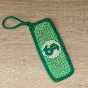 Könnte beinhalten: Handgefertigter, grüner Häkel-Lesezeichen in Form eines Geldscheins. Es hat einen weißen Kreis mit einem grünen Dollarzeichen und eine grüne Schlaufe oben.