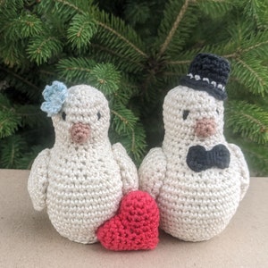 May include: Two crocheted white birds, one wearing a black top hat and bow tie, the other with a blue flower, holding a red heart.