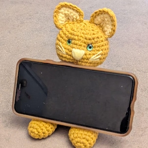 Puede incluir: Un soporte para teléfono de gato de ganchillo amarillo hecho a mano. El gato tiene orejas de color amarillo claro, ojos verdes y bigotes blancos. El teléfono se sujeta en una funda marrón. El soporte para teléfono está sobre una superficie gris.