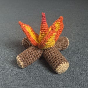 Könnte beinhalten: Ein gehäkeltes Lagerfeuer-Spielzeug mit braunen Holzscheiten und orangefarbenen und gelben Flammen.