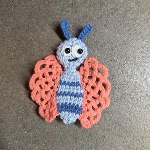 May include: A crocheted butterfly applique featuring a light blue head and body with dark blue horizontal stripes. The butterfly has two blue antennae, two black bead eyes, a stitched mouth, and two large, lacy, coral-colored wings.