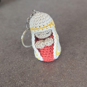 Op de afbeelding: Een gehaakte sleutelhanger met een klein figuurtje met gesloten ogen en gevouwen handen in gebed. Het figuurtje draagt een rode jurk met een witte en gele gehaakte mantel.