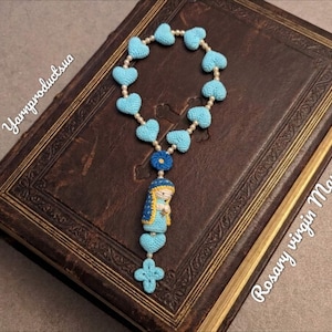 Peut inclure: Un chapelet fait à la main avec des perles en forme de cœur bleu clair et un pendentif Vierge Marie. Le chapelet est présenté sur un livre ancien avec des pages dorées. Le texte "Rosary virgin Mary" est écrit sur l'image.