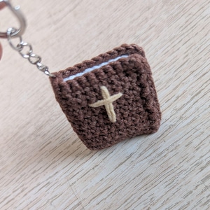 May include: A crocheted brown book-shaped keychain with a light tan cross on the front. The keychain has a silver metal ring and chain. The pages of the book are white.