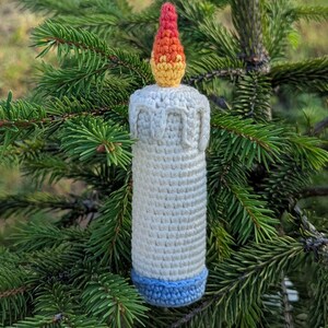 May include: A crocheted candle decoration with a white body, a blue base, and a flame in yellow, orange, and red. The candle is set against a backdrop of green pine needles, suitable for Christmas or festive occasions.