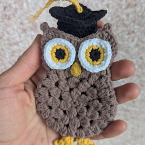 May include: A brown crocheted owl with yellow feet and a black graduation cap. The owl has white eyes with yellow and black accents.