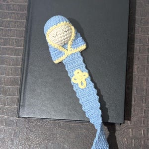 May include: A handmade crochet bookmark featuring a sleeping baby design. The bookmark is primarily light blue with yellow trim and a yellow flower accent. It rests on a black book, perfect for book lovers.