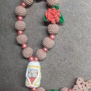 May include: A crocheted rosary with pink and brown beads, a small crocheted figure of a baby, and two crocheted flowers with green leaves.