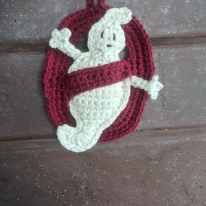 Könnte beinhalten: Ein gehäkeltes Ornament mit einer cremefarbenen Geisterfigur, die in einem dunkelroten ovalen Ring zentriert ist. Der Geist hat zwei nach außen gestreckte Arme und das gesamte Stück enthält eine kleine gehäkelte Schlaufe zum Aufhängen.