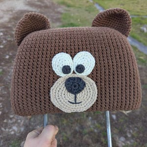 Puede incluir: Una funda de reposacabezas de oso marrón tejido a ganchillo con una cara sonriente, ojos blancos y pupilas gris oscuro. El oso tiene dos orejas marrones y está sujeto a varillas de soporte metálicas. El reposacabezas está diseñado para encajar sobre un reposacabezas de asiento de coche.