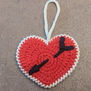 May include: A handmade, red crocheted heart-shaped ornament with a white scalloped border and a white loop for hanging. Two black arrows are stitched across the heart. The ornament is approximately 4 inches wide.