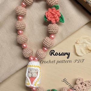 May include: A rosary crochet pattern PDF featuring a string of beige crocheted spheres interspersed with small pink beads. The design includes two coral-colored crocheted flowers with green leaves and a central white, yellow, and coral figure.