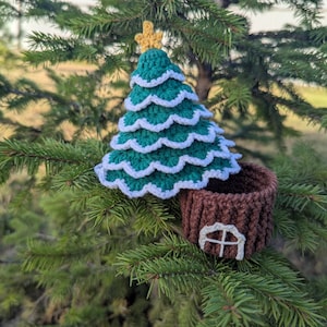 Puede incluir: Adorno navideño hecho a mano con ganchillo, con un árbol verde y ribete blanco, coronado por una estrella dorada. Debajo, un soporte marrón con forma de tocón de árbol con una ventana blanca. El adorno se muestra en una rama de pino verde.