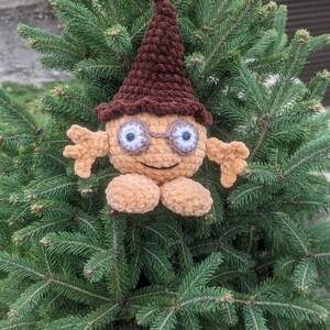 Pode incluir: Um brinquedo de pelúcia de crochê com um chapéu marrom e olhos grandes e brancos. O brinquedo está sentado em uma árvore de abeto verde.
