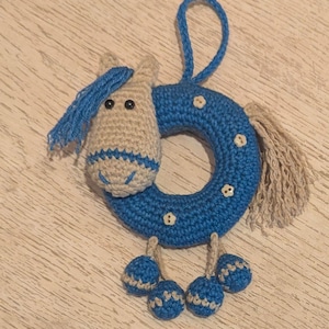 May include: A handmade crochet horse-shaped baby rattle. The horse head is beige with blue accents and black eyes. The blue ring has white flower accents and dangling blue and beige balls. A blue loop is attached for hanging.