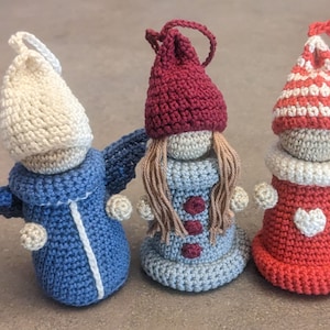 Può includere: Tre ornamenti all'uncinetto a forma di persone che indossano cappelli. L'ornamento a sinistra indossa un cappello bianco e un cappotto blu. L'ornamento al centro indossa un cappello rosso e un vestito blu. L'ornamento a destra indossa un cappello a righe rosso e bianco e un vestito rosso con un cuore bianco.
