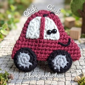 May include: A burgundy crochet car with white accents, black wheels, and black button eyes. The car has the words "Click Clack" above it and "Yarnproductsua" below it. The wheels have gray detailing. The car is sitting on a surface with green moss.