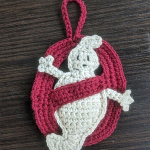 May include: Crocheted ornament featuring the Ghostbusters logo. The design includes a white ghost figure inside a red circle with a red band across the middle. A loop is attached for hanging.