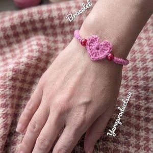 May include: A pink crochet bracelet featuring a heart-shaped design. The bracelet has red beads on either side of the heart. The word "Bracelet" is written in cursive. The bracelet is on a hand.
