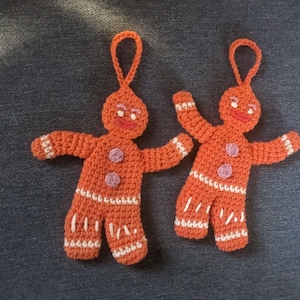 Pode incluir: Dois enfeites de gingerbread man de crochê com acabamento branco e botões rosa. Os enfeites são laranja e têm rostos sorridentes.