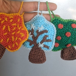 May include: Three crocheted trees, each representing a different season. The first tree is orange and yellow with brown leaves, the second tree is blue with a brown trunk and white snowflakes, and the third tree is green with red apples.